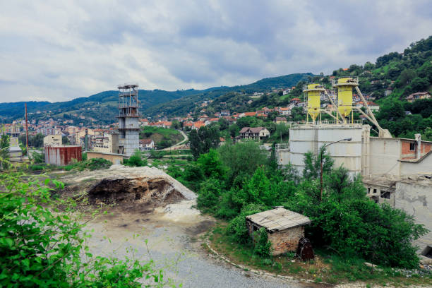 An industrial site is visible in Kosovo, surrounded by lush greenery and hills, with construction equipment and small buildings dotting the landscape on a cloudy day.