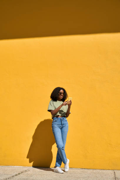 istock Young happy African girl standing at yellow wall using mobile phone. Vertical 2172556652