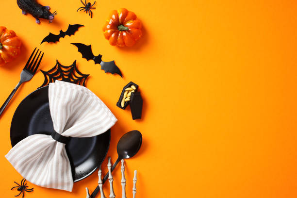 Halloween table setting with scary decorations, top view. Flat lay black plate with napkin, cutlery, bats, spiders, pumpkins, skeleton hands on orange background
