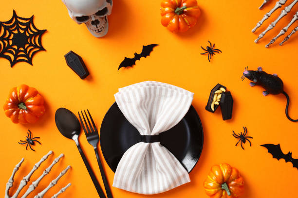 istock Halloween table setting with scary decorations. Black plate with napkin, cutlery, bats, spiders, coffin with maggots, pumpkins, skull on orange background. Flat lay, top view. 2172560487