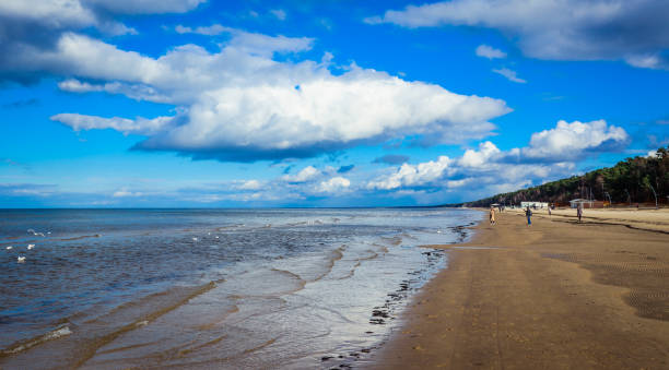 Visitors enjoy a leisurely walk on Jurmala's sandy beach, taking in the refreshing sea breeze and beautiful blue skies, perfect for a sunny day outdoors.