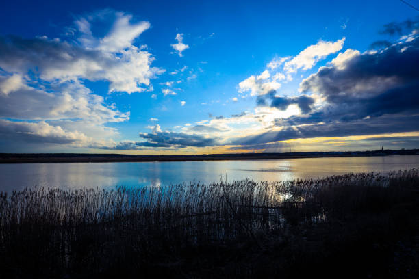 A stunning sunset bathes the Jurmala landscape in warm hues as the sun descends, casting reflections on the calm water while clouds dance across the sky.