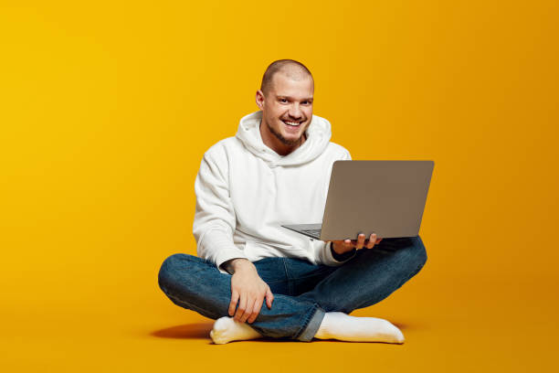 istock Happy young bearded man in white hoodie holding laptop computer while sitting on a floor 2172590837
