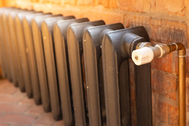 A traditional retro metallic radiator is connected to a brass pipe in a warm and inviting interior setting. The radiator features a classic design and is positioned against a rustic exposed brick wall, adding character to the room. This heating element is essential for providing warmth during winter months.