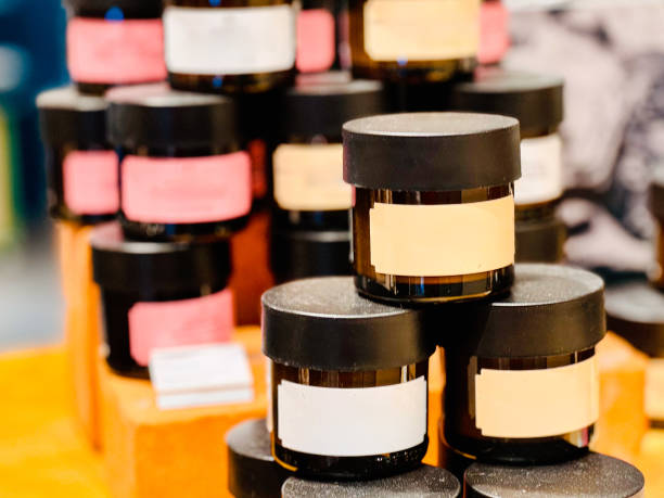 : A display of neatly stacked small cosmetic jars with black lids and blank labels, arranged on wooden blocks. The jars come in different pastel colors, indicating a variety of products.