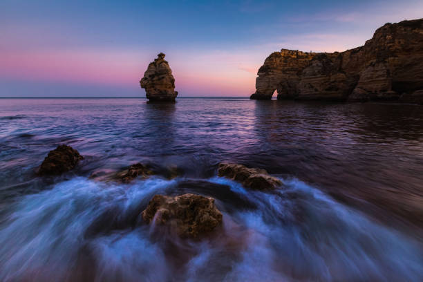 Soft hues of the evening sky reflect on the calm waters of Praia de Marinha. In the foreground, rocks with subtle shadows rest quietly in the water, while distant cliffs and an isolated rock pillar appear dreamlike under the glowing sunset.