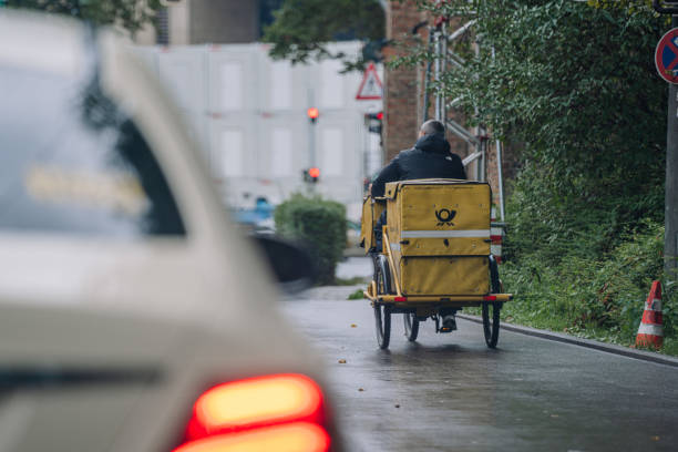 Munich, Germany - September 13, 2024 -  Postal worker riding a yellow cargo tricycle on a rainy street, with blurred car and traffic lights in the background.