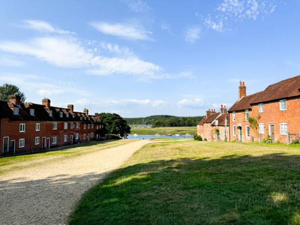 Stock photo showing rows of redbrick, terraced cottages with a gravelled sloping pathway leading down towards the Beaulieu River at Buckler's Hard, New Forest, Hampshire, England, UK. Now a popular tourism spot the hamlet of Buckler's Hard was once renown for shipbuilding. imagen de ladrillo rojo, cabañas en terrazas y camino inclinado de grava que conduce al río beaulieu, buckler's hard, new forest, hampshire, inglaterra, reino unido, hileras de casas y zona de hierba, barcos y yates amarrados en el agua, cielo azul nubla