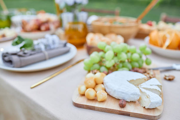 Serving cheese platter with fresh grapes and assorted appetizers on wooden board placed on table outdoors during daylight gathering