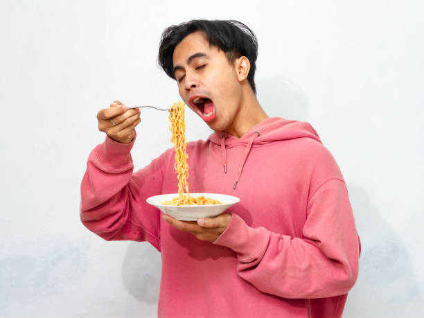 istock Young Asian man in a pink hoodie showing a joyful and euphoric expression while eating a plate of instant fried noodles, isolated on a white background. 2172752524