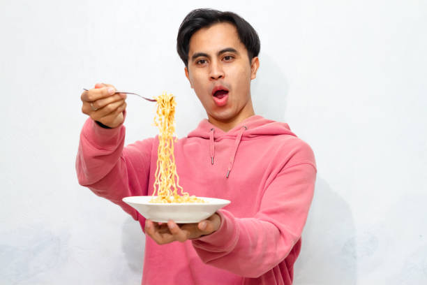 istock Young Asian man in a pink hoodie showing a joyful and euphoric expression while eating a plate of instant fried noodles, isolated on a white background. 2172752579