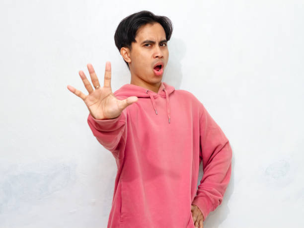 istock Young Asian man in pink hoodie with an angry expression, posing with a stop gesture using his hand. 2172756202