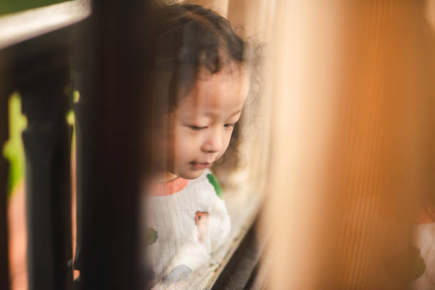 A young girl stands quietly by a window, framed by sheer curtains that softly filter the morning light into the room. Dressed in a colorful, patterned sweater, she gazes contemplatively, with her hand resting on her neck, capturing a serene and introspective moment. The softly lit scene, with the blurred greenery visible through the window, creates a calm and cozy atmosphere, evoking themes of childhood, solitude, and the quiet beauty of everyday moments.