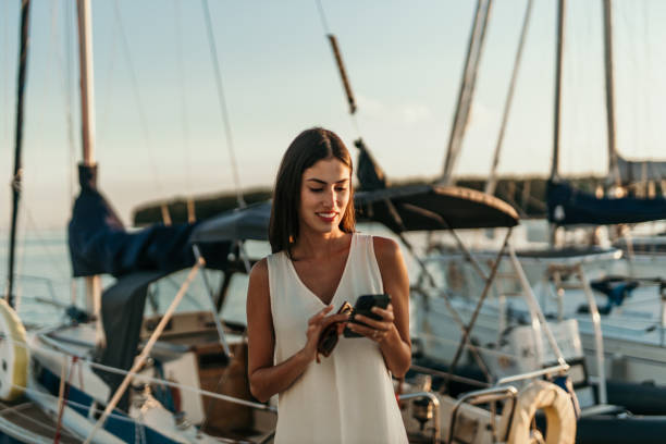 Golden hour walk and phone chat Happy rich female traveler using a smartphone at the marina on vacation. mediterranean woman portrait stock pictures, royalty-free photos & images