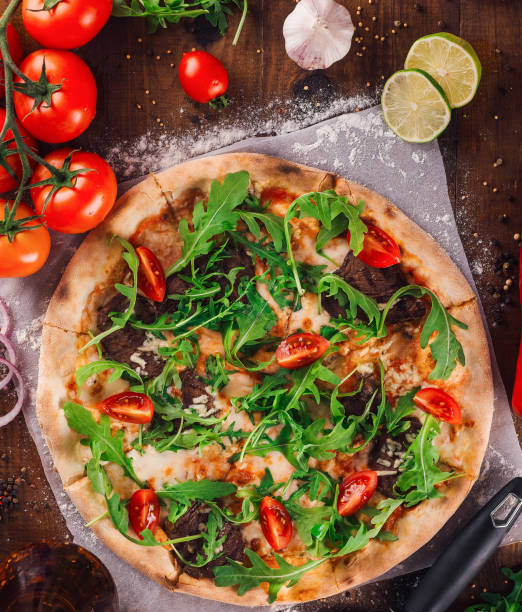 Delicious pizza with arugula, tomatoes and meat freshly baked and served on wooden table surrounded by ingredients