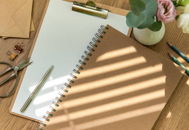A workspace with stationery including a notepad, clipboard, and scissors next to a vase of flowers in soft afternoon light