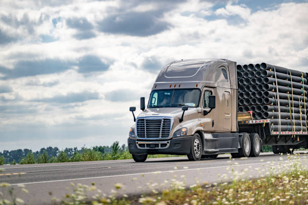 Beige big rig semi truck transporting fastened plastic pipes on step deck semi trailer running on the summer highway road going between meadows Industrial long hauler carrier beige big rig semi truck with step deck semi trailer transporting fastened plastic pipes running on the summer highway road going between meadows truck in rain stock pictures, royalty-free photos & images