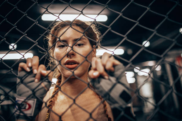 A determined female boxer peers through a chain link fence, exemplifying focus and strength, while preparing mentally and physically for the training ahead.