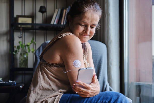 medium shot of a confident caucasian middle-aged female with type 1 Diabetes using an smartphone app to check her glucose levels through a glucose sensor during the day at home stock photo