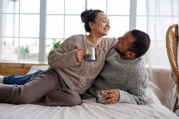 istock Cheerful young multiethnic couple in sweaters holding coffee while hugging and relaxing on bed at home 2172805058