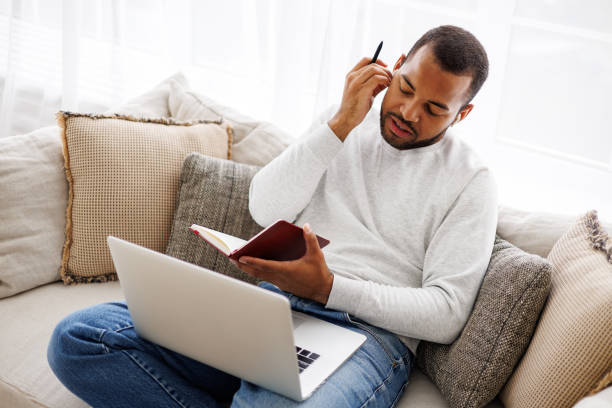 istock Young african american student using earphones while holding notebook near laptop on couch at home 2172805506