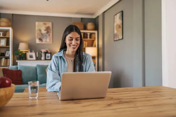 A young cheerful woman sitting at the table in the living room at home and using a laptop. A young cheerful woman sitting at the table in the living room at home and using a laptop.
