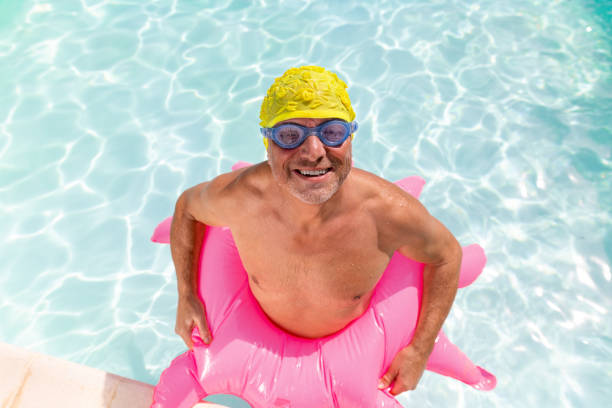 Medium portrait shot of a senior man standing in a swimming pool outside a house in Castelferrus near Toulouse - France. He is wearing a yellow flower swimming cap and has blue swimming goggles on his face. He is looking and smiling at the camera and has a pink inflatable ring around him.\n\nVideos are available similar to this scenario.