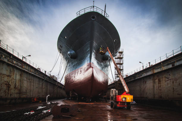 A dramatic low-angle shot of the massive bow of a bulk carrier in drydock, showcasing the imposing scale of the vessel as it towers above. The ship’s hull is exposed, with scaffolding, a cherry picker and maintenance equipment visible around the drydock area. The anchor and anchor chain of the vessel lie in front of it. A team of workers is painting the hull under a dramatic cloudy sky. The image captures the engineering marvel of large-scale shipbuilding and repair, ideal for themes of maritime industry, ship maintenance, heavy machinery, and industrial work
