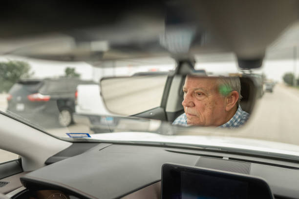 Reflection of senior man sits in driver's seat, visible in rear-view mirror on sunny day. The atmosphere feels peaceful and relaxed, capturing a moment of quiet contemplation during the drive.