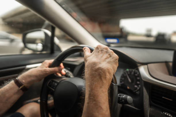 Cropped hands of senior man grip steering wheel while driving car from point of view perspective. The hands show steady control and confidence on the road.