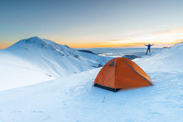 Hiker with open arms standing in front of the tent on snowy mountain admiring sunset, Alps, Italy