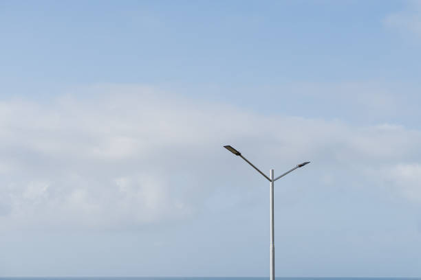 A solitary street lamp stands against a clear blue sky with soft clouds. The lamp features a modern design with two light fixtures extending from a tall pole, overlooking a tranquil sea.