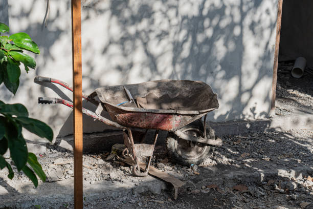 A weathered red wheelbarrow sits on a gravel surface, partially shaded by trees. The wheelbarrow is filled with dirt and tools, with a wall in the background casting soft shadows.