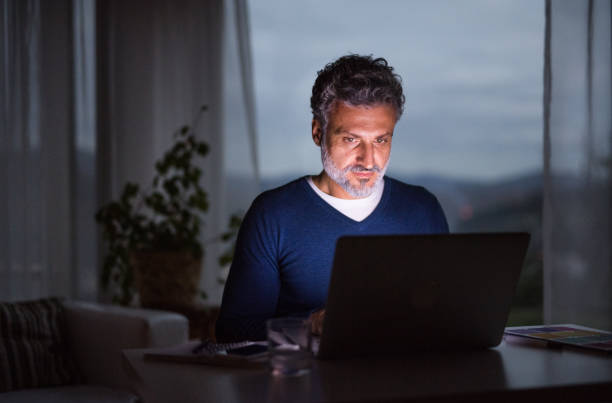 istock Handsome man working from home office at evening, using laptop. Screen glow illuminates man's face. Telecommuting and home office. 2172902110