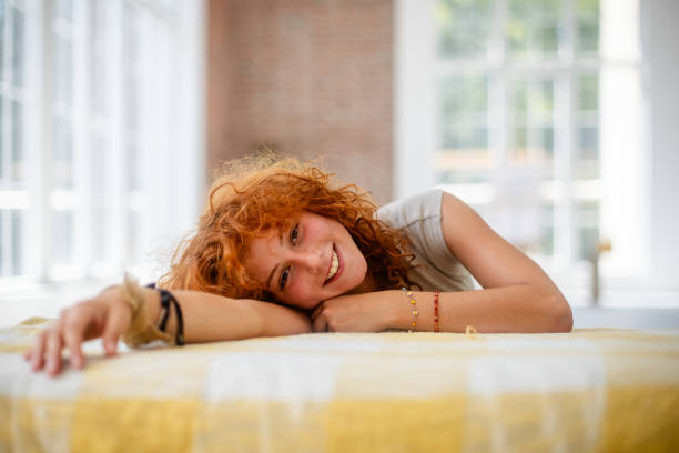 A red-haired woman lies on a couch in a sunlit loft with large windows and a brick wall, her casual attire and relaxed position reflecting tranquility and modern comfort.