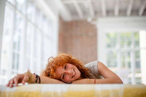 A woman with curly red hair rests her head on a table in a bright room with large windows and natural sunlight, showcasing a relaxed pose ideal for lifestyle themes.