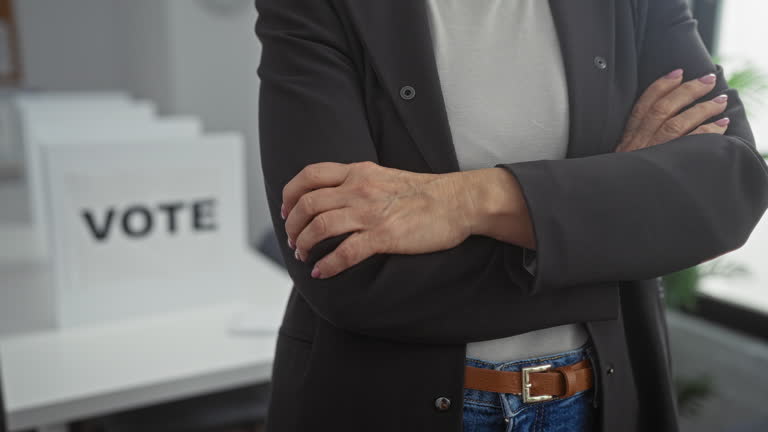 Mature woman standing with folded arms in an electoral college room surrounded by voting booths.