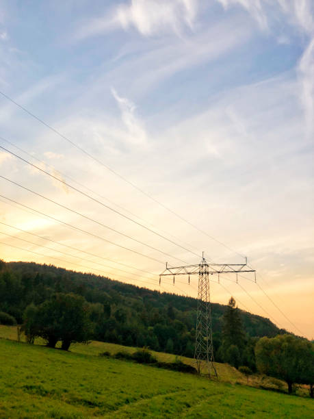 rural landscape at sunset, with a towering electricity pylon standing prominently in the middle of a lush, green field. Power lines stretch across the frame, disappearing into the distance, highlighting the intersection of nature and technology. Rolling hills covered in dense trees surround the area, with soft orange and pink hues blending into the blue sky as daylight fades. The serene natural setting contrasts with the industrial presence of the power infrastructure, making it an ideal image for themes related to energy, nature, or rural development