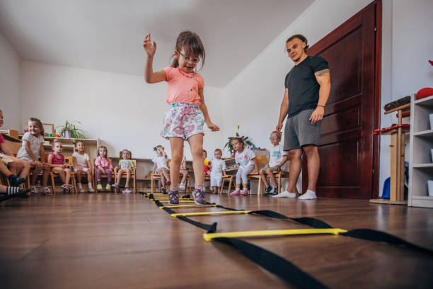 A young girl participates in a classroom activity under the supervision of a teacher, with classmates seated in the background. The activity fosters physical coordination and classroom engagement.
