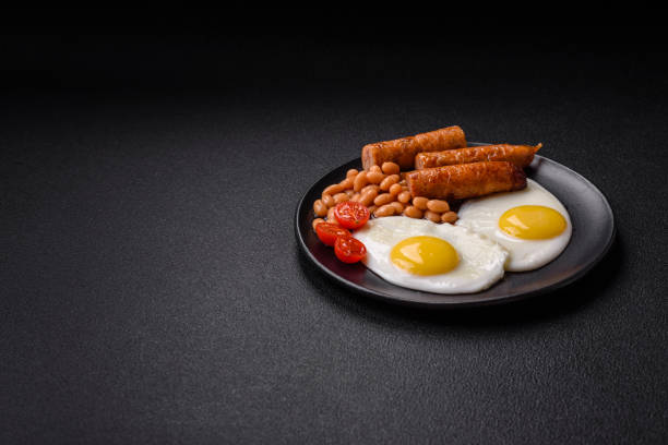 Full English breakfast with boiled eggs, beans, sausages, tomatoes, salt and spices on dark concrete background