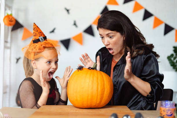 istock A mother and daughter, both dressed for Halloween, react with playful surprise as they look at a plastic spider on a large pumpkin. The scene is festive with Halloween decorations in the background. 2172948110