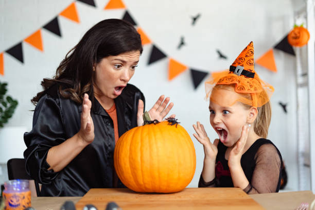 istock A mother and daughter, both dressed for Halloween, react with playful surprise as they look at a plastic spider on a large pumpkin. The scene is festive with Halloween decorations in the background. 2172948369