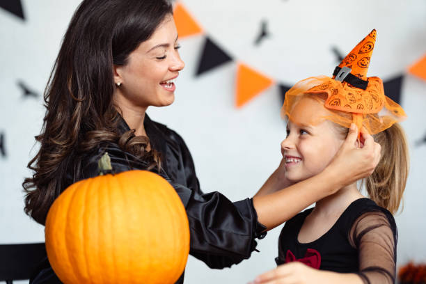 istock A mother helps her daughter adjust a cute Halloween witch hat while they smile at each other. A carved pumpkin sits on the table, and festive Halloween decorations hang in the background. 2172948493