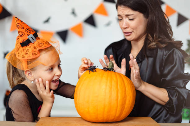 istock A mother and daughter, both dressed for Halloween, react with playful surprise as they look at a plastic spider on a large pumpkin. The scene is festive with Halloween decorations in the background. 2172948562