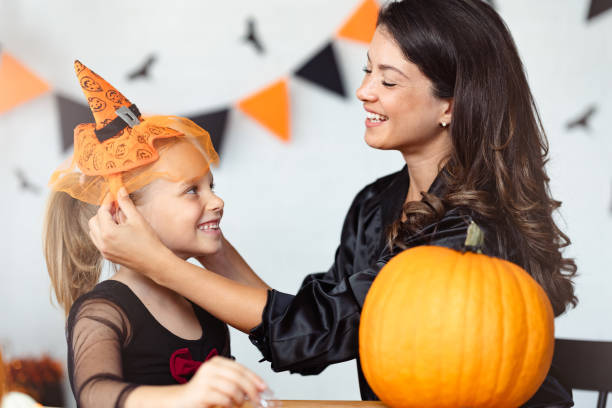 istock A mother helps her daughter adjust a cute Halloween witch hat while they smile at each other. A carved pumpkin sits on the table, and festive Halloween decorations hang in the background. 2172949477