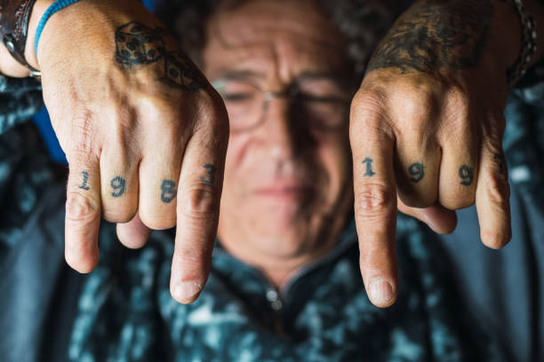 Blurred unrecognizable middle aged male with curly hair and tattoos on hands without a thumb in trendy outfit and eyeglasses showing rock n roll gesture against blue background