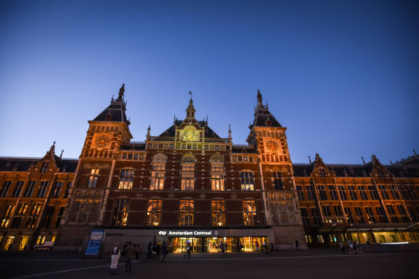 Amsterdam, Netherlands - July 1, 2018: This picture captures the iconic Amsterdam Centraal, the central railway station of Amsterdam, Netherlands. The photograph was taken during twilight, with the sky transitioning from day to night, creating contrast between the deep blue sky and the warm, illuminated façade of the building. The architectural style is a blend of Gothic and Renaissance Revival, with intricate details, tall towers, and a clock at the center.