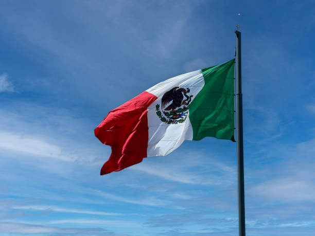 Mexico flag hangs on a pole, blowing in the wind. The sky is cloudy with partial blue.