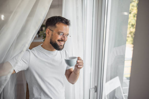 istock A young man enjoys his morning coffee while standing by the window and looking out into his yard 2173050007