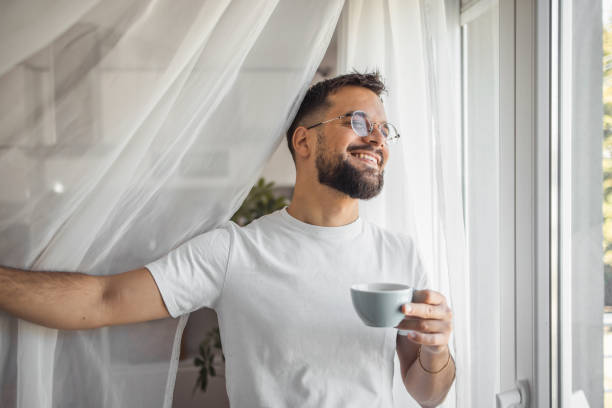 istock A young man enjoys his morning coffee while standing by the window and looking out into his yard 2173050008